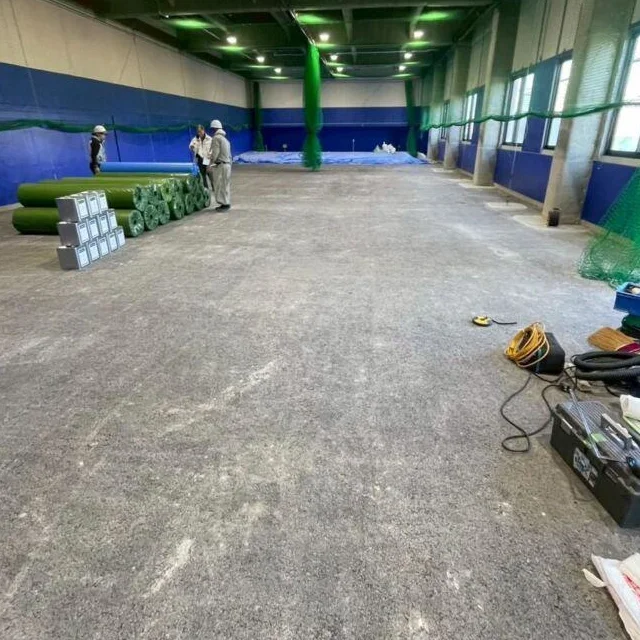 青い壁と防球ネットのある屋内ゴルフ施設で、ロール状の人工芝を運び込み施工を進めているスタッフ
