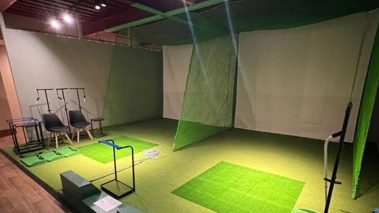 打席用の人工芝とネットを備えた屋内ゴルフ練習施設。椅子とクラブスタンドが並ぶ打席の様子